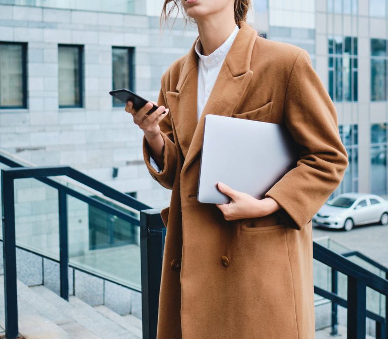 Young lawyer in coat with laptop and cellphone thoughtfully looking away outdoor